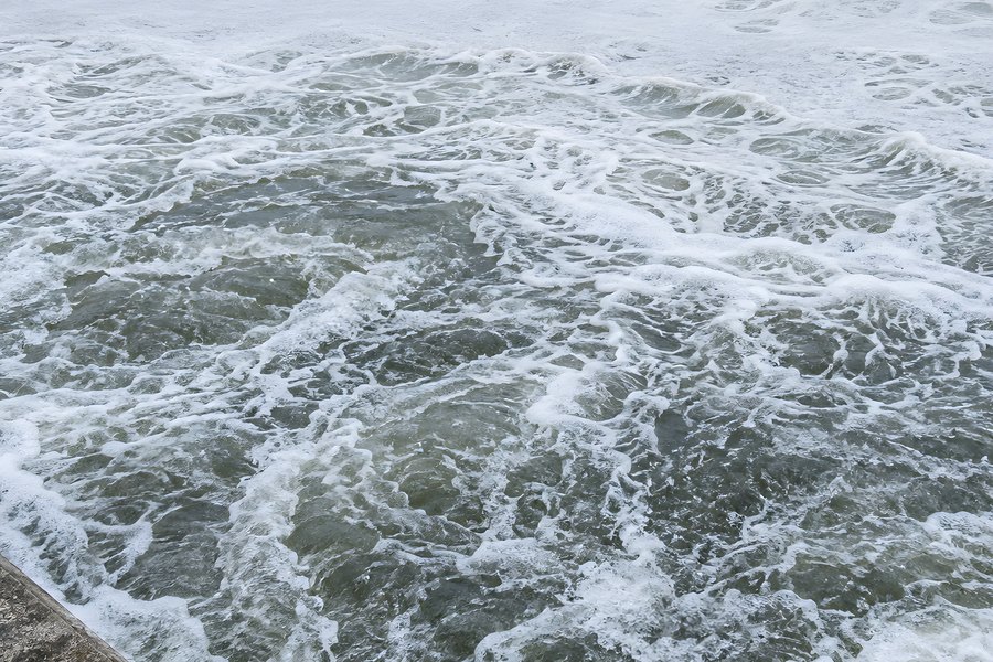 Turbulent foaming water churns against a diagonal concrete weir edge in close-up aerial view