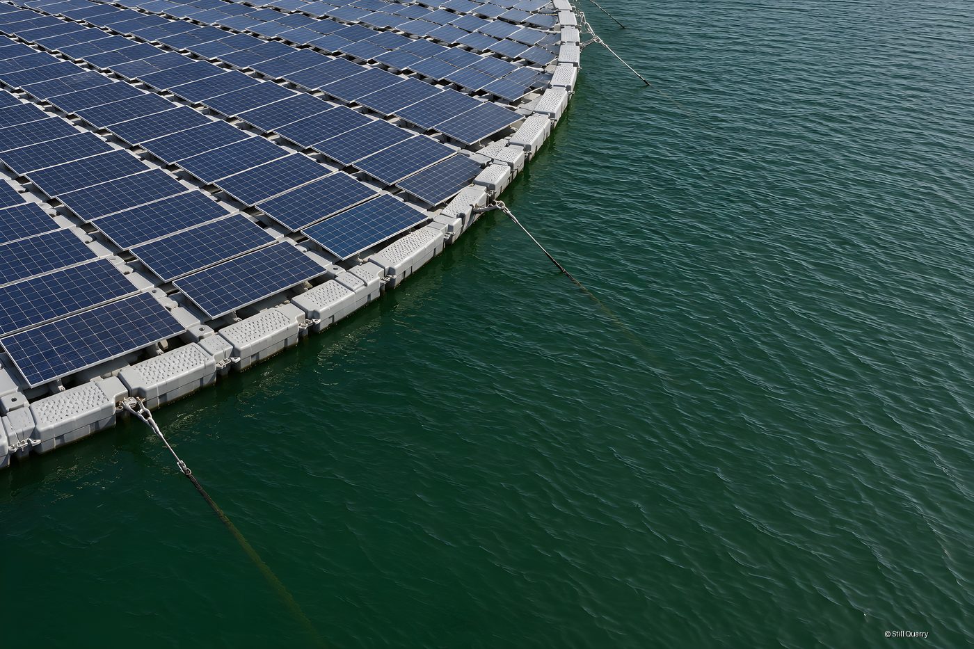 High angle view of a floating solar farm's curved edge, grey pontoon floats and mooring cables on rippled green reservoir water