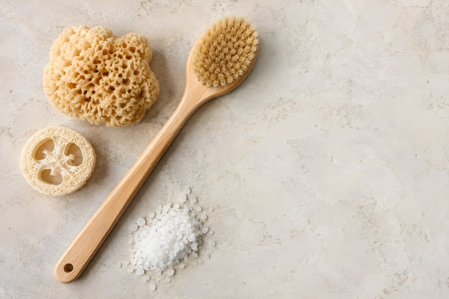 Wooden dry body brush, natural sea sponge, loofah slice and sea salt on marble background, overhead flat lay with copy space