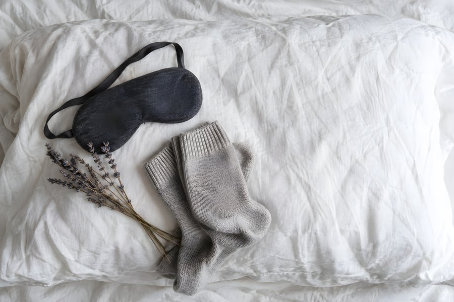 Black sleep mask, grey wool socks and a sprig of dried lavender arranged on a rumpled white pillow, top view, no people